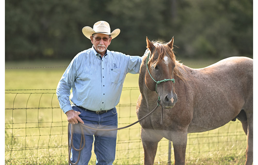 An older man in a cowboy hat and blue button-down shirt stands beside a roan horse in front of a wire fence.